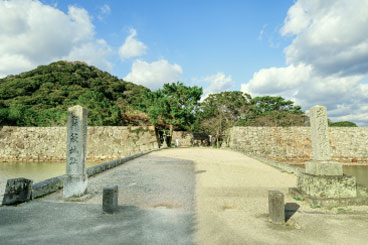 青空の下、石橋と広い道が続く風景。奥に丘が見える。