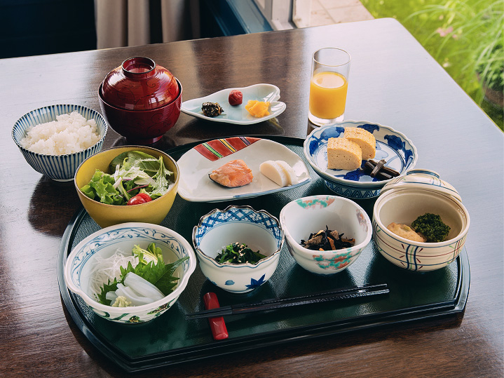 黒いお盆に並んだ旅館の朝食。白米、味噌汁、焼き魚、卵焼き、漬物、煮物、おひたし、サラダ、刺身、オレンジジュース。