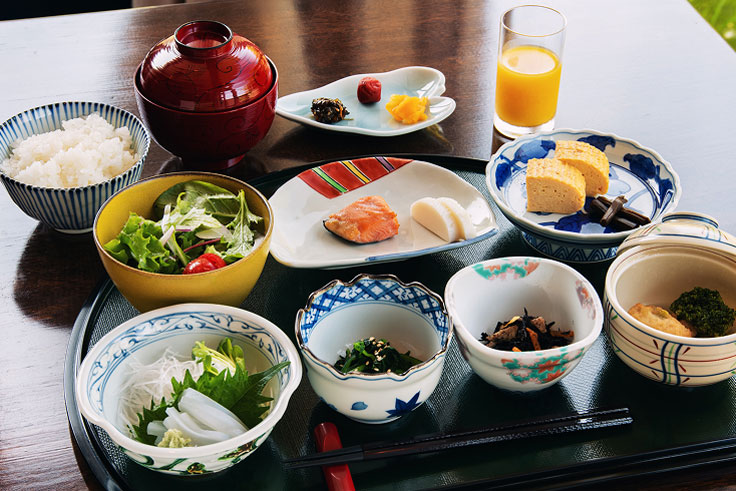 Ryokan breakfast lined up on a black tray. White rice, miso soup, grilled fish, omelet, pickles, simmered vegetables, boiled vegetables, salad, sashimi, and orange juice.