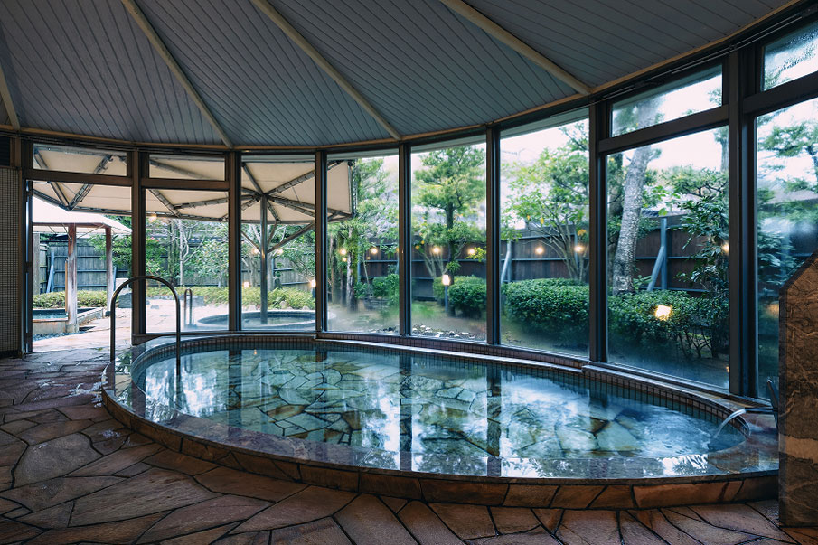A large bath surrounded by large windows overlooking the greenery of the garden. It is an open space with a stone circular bathtub and an open-air bath in the back.