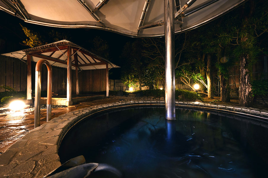 An open-air bath illuminated by lights. The surface of the water shimmers quietly, and the surrounding trees and pavilion are dimly illuminated.