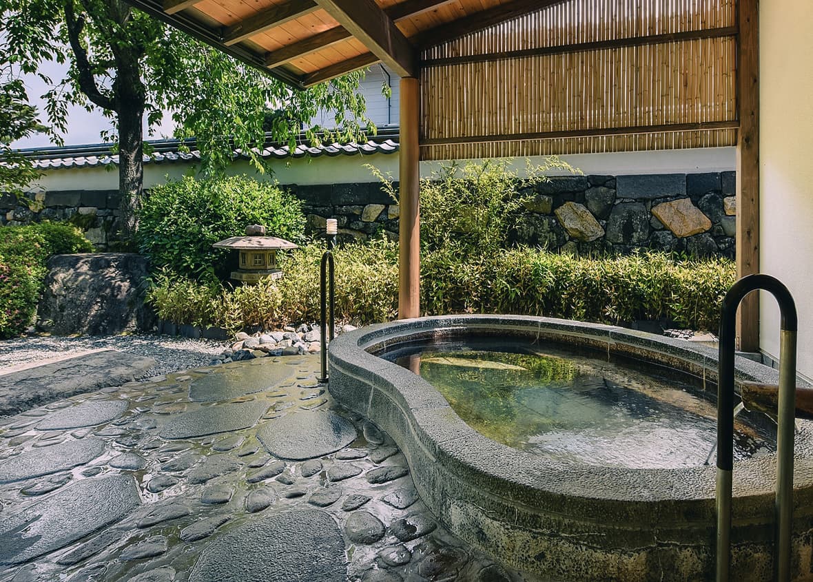 木製の屋根に覆われた、曲線を描くグレーの石造りの露天風呂。湯面は穏やかで、周囲には丸い石が敷き詰められた洗い場と、緑豊かな庭園が広がる。