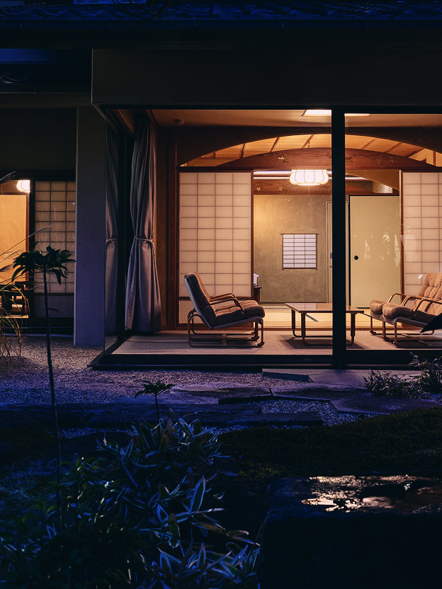 和の趣を感じる照明に包まれた、夜の客室の様子。障子や畳、座椅子が並び、外の庭園越しに室内が見える。