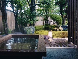 露天風呂から見える庭の風景。湯気が立ち上る木製の四角い浴槽の手前には濡れた石畳が広がり、奥には緑豊かな木々や苔、岩が配置された庭園が見えます。